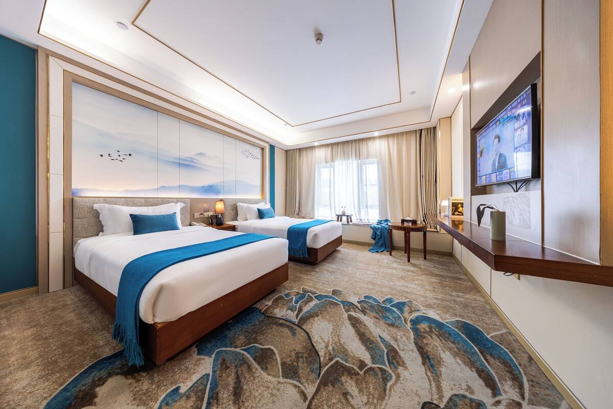 Habitación de hotel con dos camas, un televisor de pantalla plana grande, un escritorio y un mural decorativo con un paisaje.