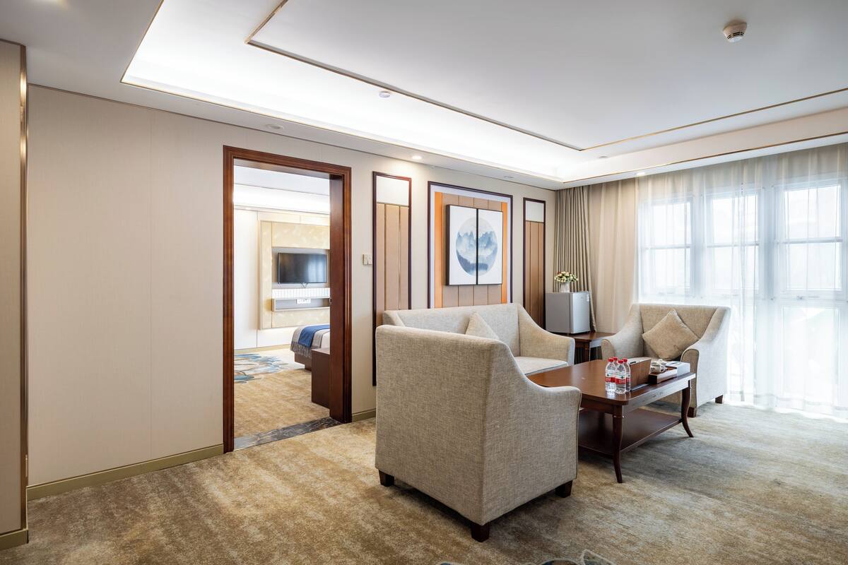 Habitación de hotel con sofá, sillones, una mesa de centro y televisión. Se ve una puerta que da al ba ño y a un pasillo.