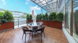 A covered outdoor area with a glass table and wicker chairs, surrounded by wooden planters and a view of a building in the background. - Xinrun Hua Hotel Hongqiao Park Branch (Shenzhen)