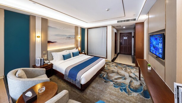 A hotel room with a large bed, a desk, a chair, a TV, and a view of the hallway. - Xinrun Hua Hotel Hongqiao Park Branch (Shenzhen)