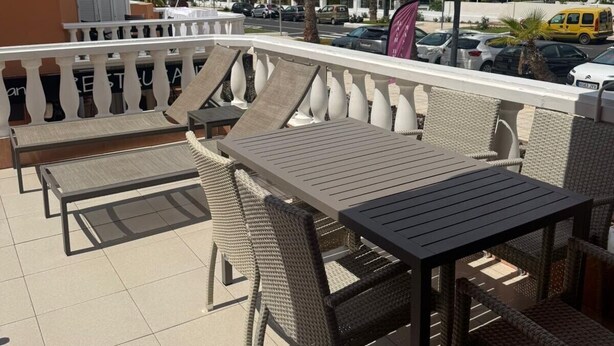 An outdoor seating area with wicker furniture and a white railing, overlooking a street with palm trees and parked cars.