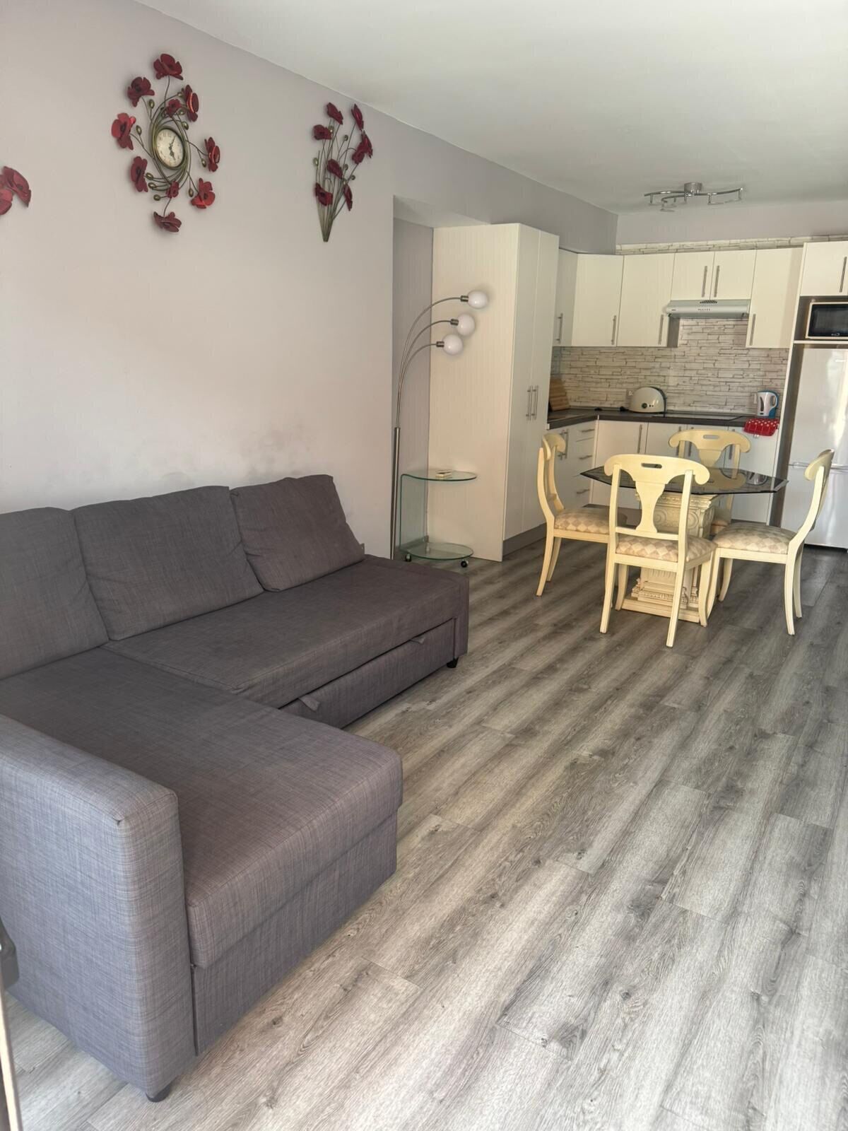 Un salon moderne avec un canapé d’angle gris, un coin repas avec une table en verre et des chaises en bois, et une cuisine avec des meubles blancs et un réfrigérateur en acier inoxydable.