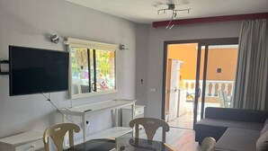A living room with a glass-top dining table, wooden chairs, a sofa, a flat-screen TV, and a balcony with a view of the outdoors.