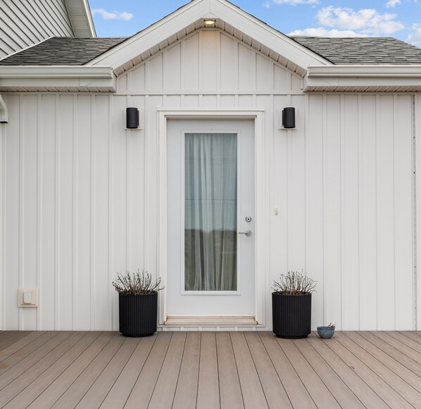 A white building with a door, two black planters with dried plants, and a wooden deck. - Large PEI Home with Bunk Room, Two Kitchens & Relaxing Outdoor Deck (Prince Edward Island)