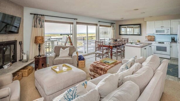 Amplia sala de estar con chimenea, un conjunto de sofás, una mesa de comedor y una zona de cocina con armarios blancos.