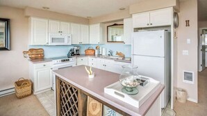 Una cocina con gabinetes blancos, microondas, refrigerador y una isla central con un tazón de vidrio.