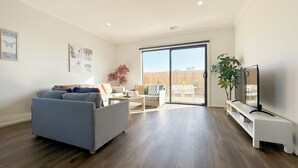 A spacious living room with a grey sofa, a coffee table, and a TV on a stand. There is a sliding glass door leading to a patio area with outdoor furniture.