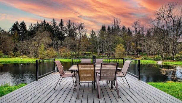 Une terrasse en bois avec une table et des chaises donnant sur un étang, entourée d’arbres et illuminée par le ciel au coucher du soleil.