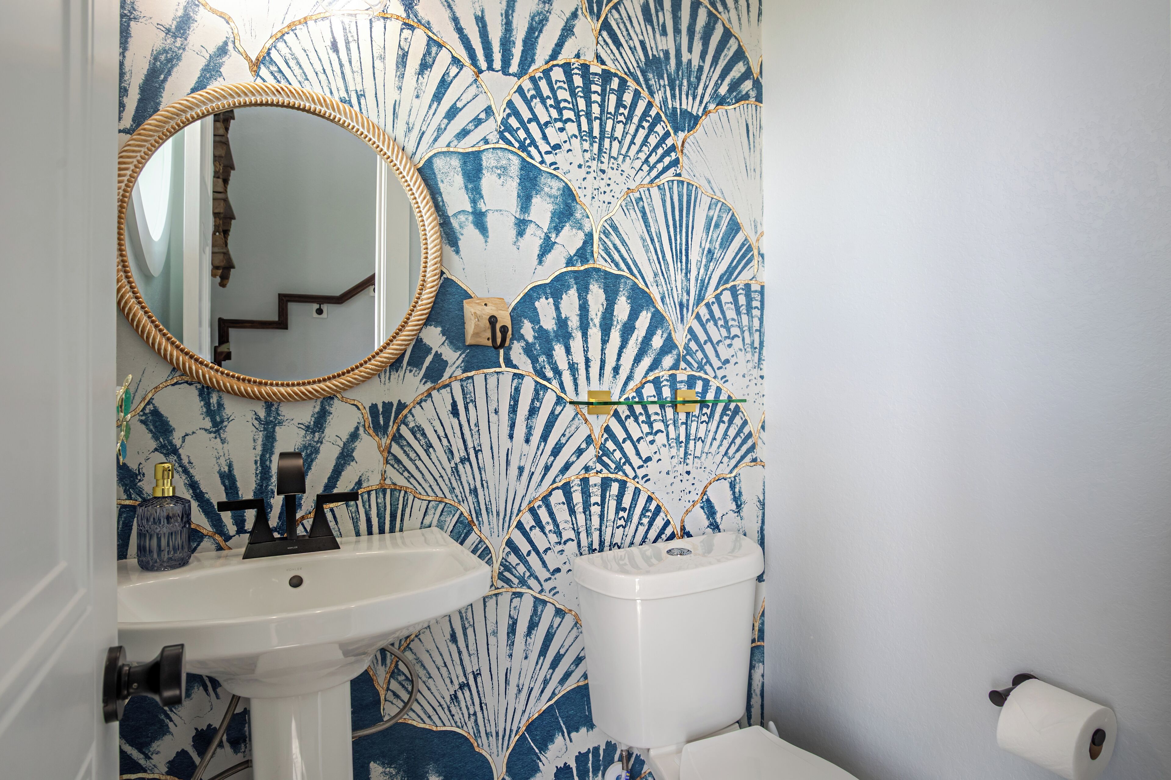 Ein Badezimmer mit einem weißen Waschbecken, einem runden Spiegel und einer Toilette. Die Wand ist mit einem blau-weißen Muschelmuster versehen.