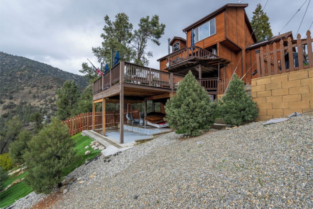 A two-story wooden cabin with a balcony, surrounded by trees and a mountainous landscape.