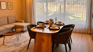 Comedor con una mesa redonda de madera dispuesta para cuatro personas, rodeada de sillas. Se observa un sofá y una mesita de centro al fondo.