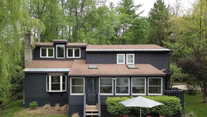 Une maison à deux étages dotée d’un patio couvert, d’une petite terrasse et d’un ensemble de chaises Adirondack rouges.