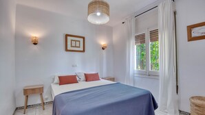A bedroom with a bed, two bedside tables, a hanging light fixture, a window with curtains, and a framed picture on the wall.