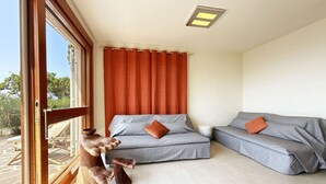 Une pièce avec deux canapés gris, dont l’un est agrémenté d’un coussin rouge, un repose-pied en bois et une fenêtre avec des rideaux orange.