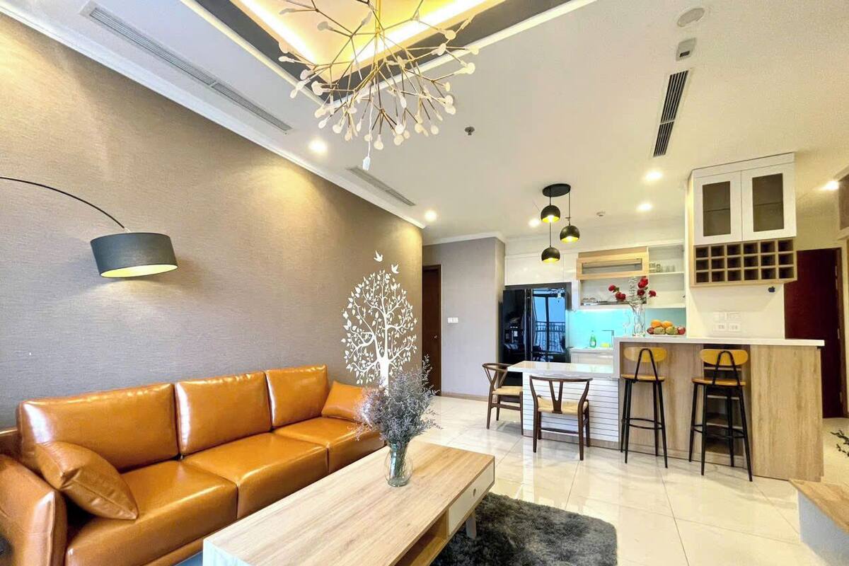Een moderne woonkamer met een bruine leren bank, een houten salontafel en een decoratieve boomsculptuur. Er is een keukenhoek met een bar en barkrukken.