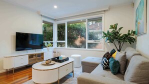 Un salon moderne doté d’un téléviseur à écran plat, d’une console blanche, d’un canapé beige et de grandes fenêtres.