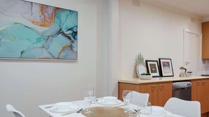 Un coin repas avec une table blanche dressée pour quatre personnes, des chaises blanches et une cuisine en arrière-plan.