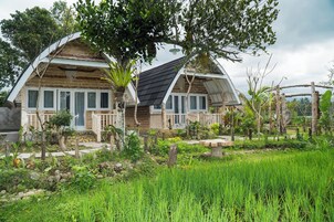Uma casa de madeira rústica com varanda, rodeada por vegetação exuberante e um campo de arroz.