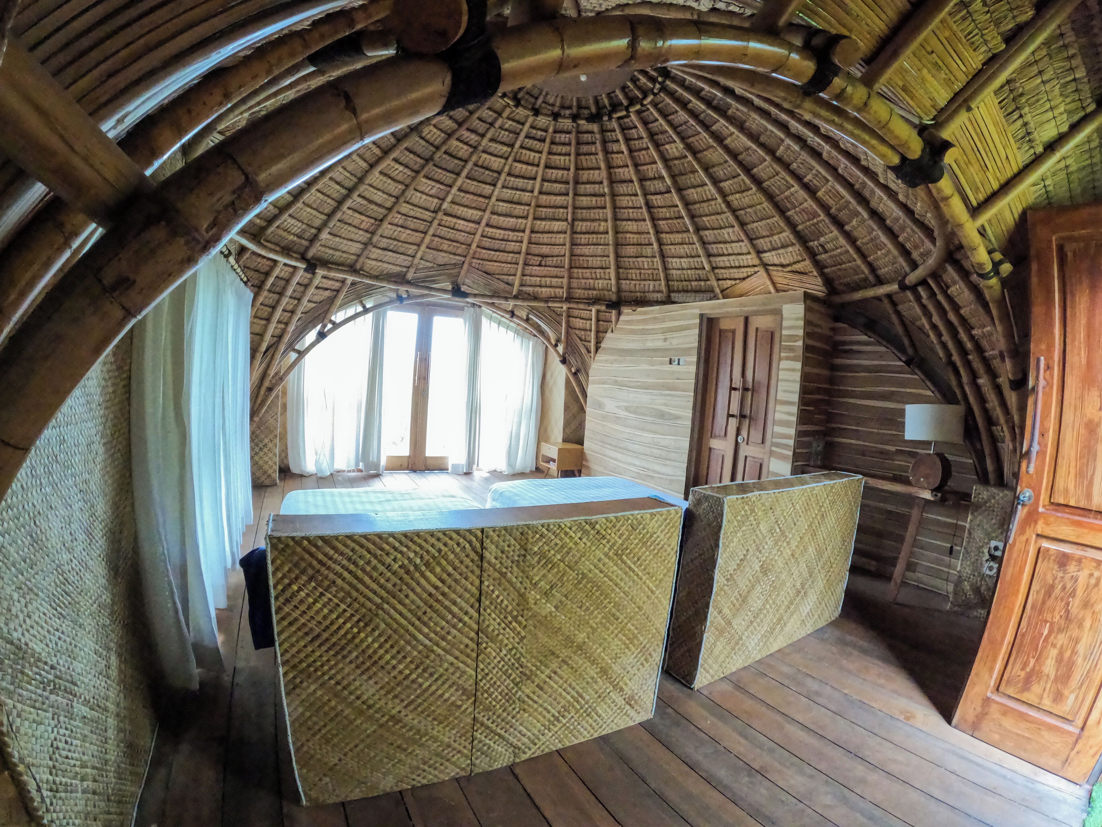 A room with a thatched roof, wooden walls, and a large window. There is a bed with a checkered bedspread and a wooden cabinet.