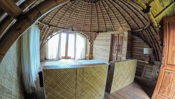 A room with a thatched roof, wooden walls, and a large window. There is a bed with a checkered bedspread and a wooden cabinet.