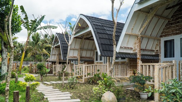 A row of traditional-style wooden houses with thatched roofs, surrounded by lush greenery and a stone pathway. - Aranka Tempasan (Pringgasela)