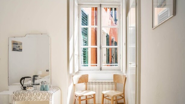 Una habitación pequeña y luminosa con ventana, un radiador, dos sillas de madera, una mesita con una tetera y un espejo.