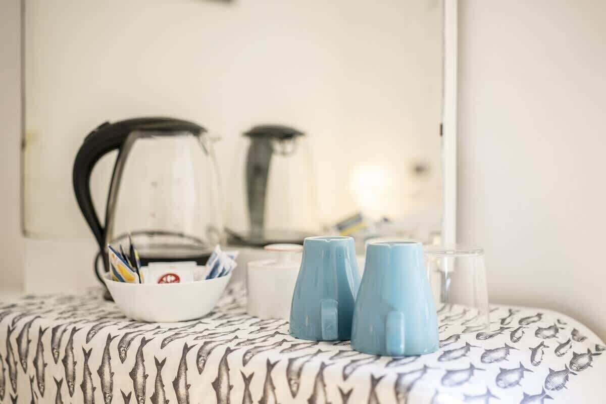 Una habitación de hotel con una colcha estampada, una cafetera, un tazón con bolígrafos y dos tazas azules sobre la mesita de noche.