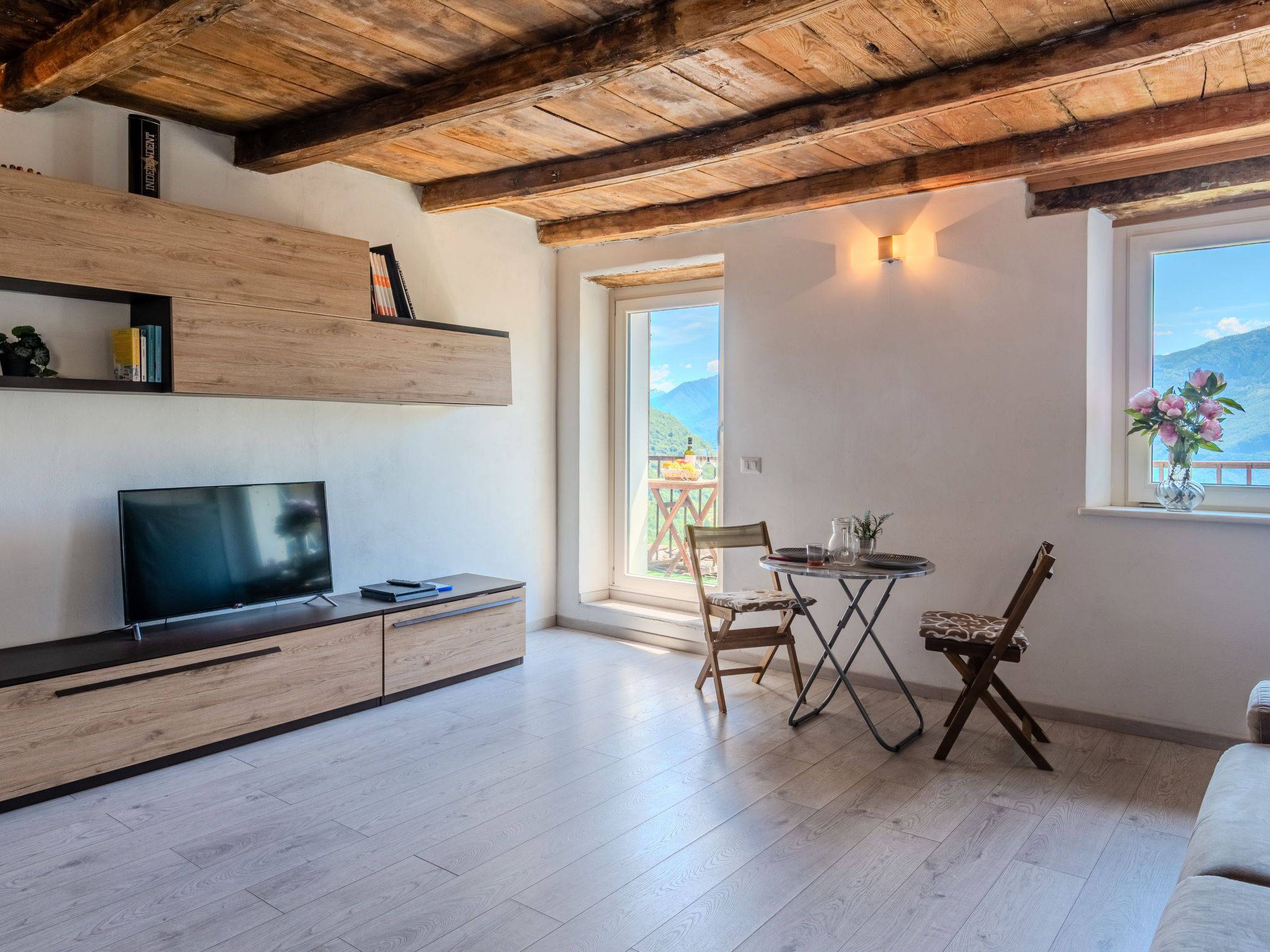 A modern living room with a TV, wooden shelves, a dining table with chairs, and a view of mountains through a large window.