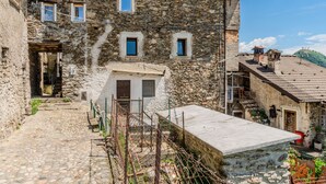 A stone building with a small entrance, a staircase leading to a door, and a fenced area with plants. - Gentile by Interhome (Gravedona)