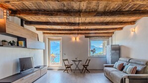 A cozy living room with a wooden ceiling, a sofa, a TV, a dining table with chairs, and a balcony with a mountain view. - Gentile by Interhome (Gravedona)