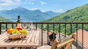 A wooden table with a bottle of wine, glasses, and a basket of fruit on a balcony overlooking a mountainous landscape. - Gentile by Interhome (Gravedona)
