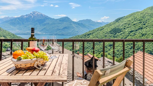 A wooden table with a bottle of wine, glasses, and a basket of fruit on a balcony overlooking a mountainous landscape. - Gentile by Interhome (Gravedona)