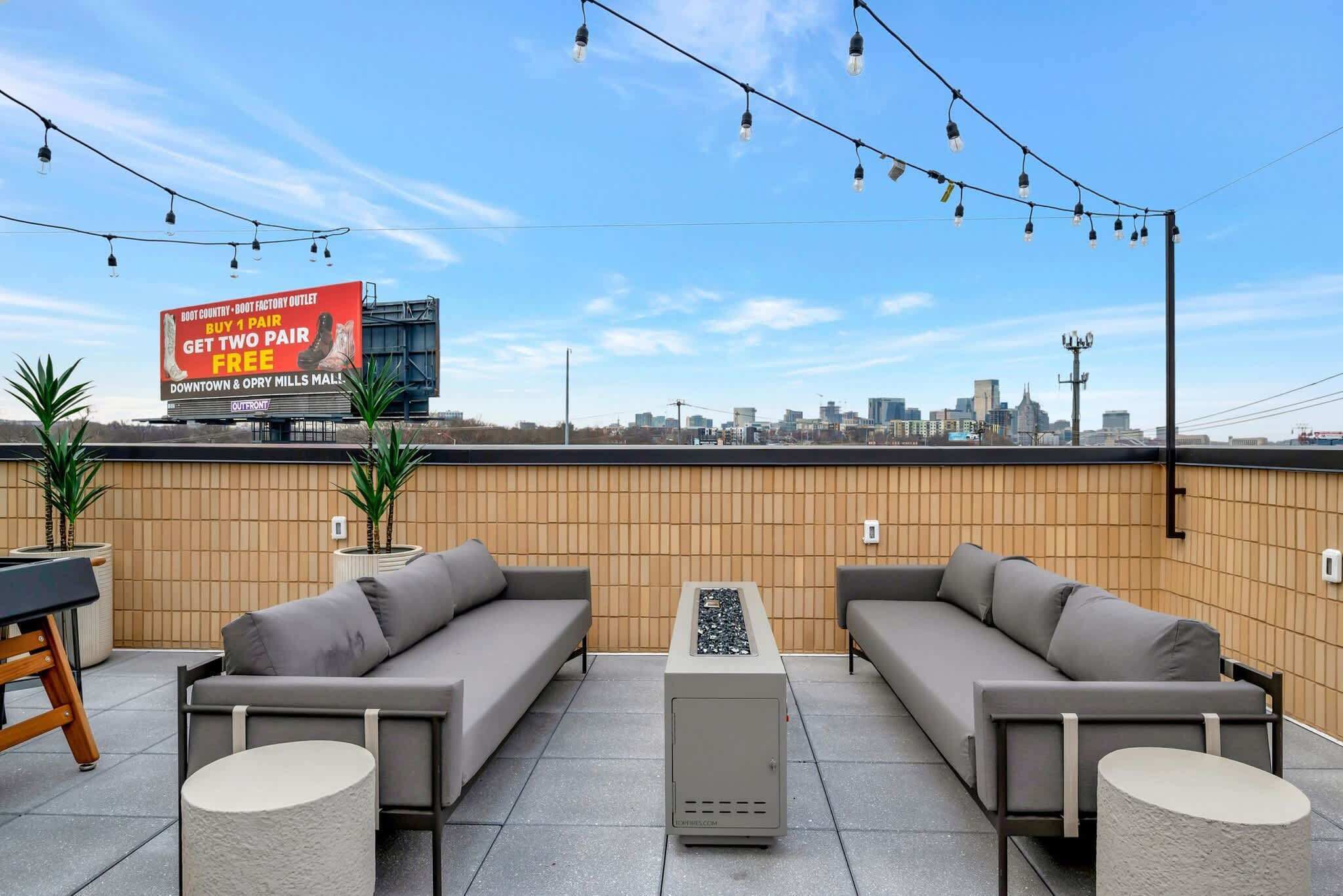 An outdoor seating area with two sofas, a small table, and potted plants. A billboard is visible in the background advertising a promotion.