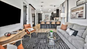 A modern living room with a grey sofa, a coffee table, and a TV. There is a dining area with a kitchen in the background. - Nashville Designer PH City Views + Rooftop Access (Nashville)
