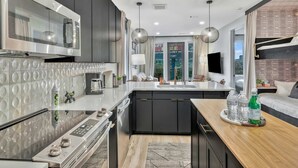 A modern kitchen with a stainless steel microwave, a built-in oven, and a large island with a sink. There are two pendant lights above the island and a flat-screen TV mounted on the wall. - Nashville Designer PH City Views + Rooftop Access (Nashville)