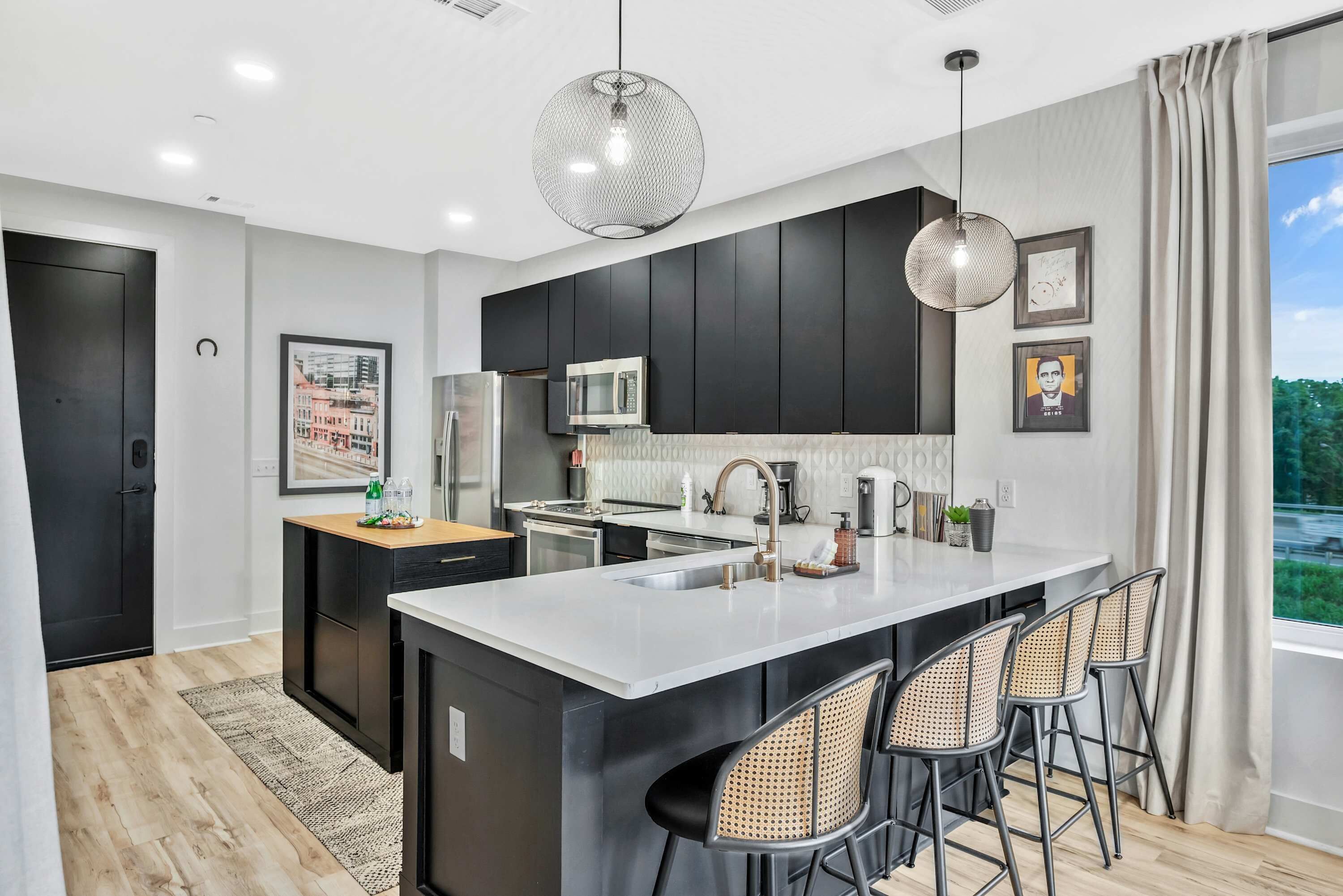 Une cuisine moderne avec des meubles de couleur sombre, un îlot central doté de tabourets, un luminaire suspendu et une grande fenêtre agrémentée de rideaux.