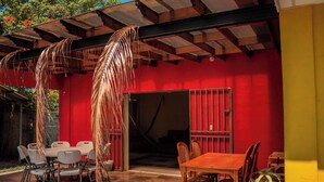 An outdoor patio with a red wall, a wooden table, and white plastic chairs. - Minerva Hotel and Restaurant (Playa San Blas)