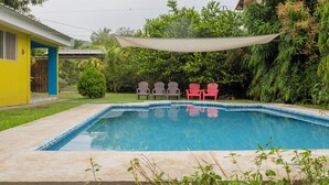 A rectangular swimming pool with a tiled edge, surrounded by a grassy area and a variety of trees. There are four Adirondack chairs, two gray and two red, positioned near the pool. A white canopy is stretched over the pool area. - Minerva Hotel and Restaurant (Playa San Blas)