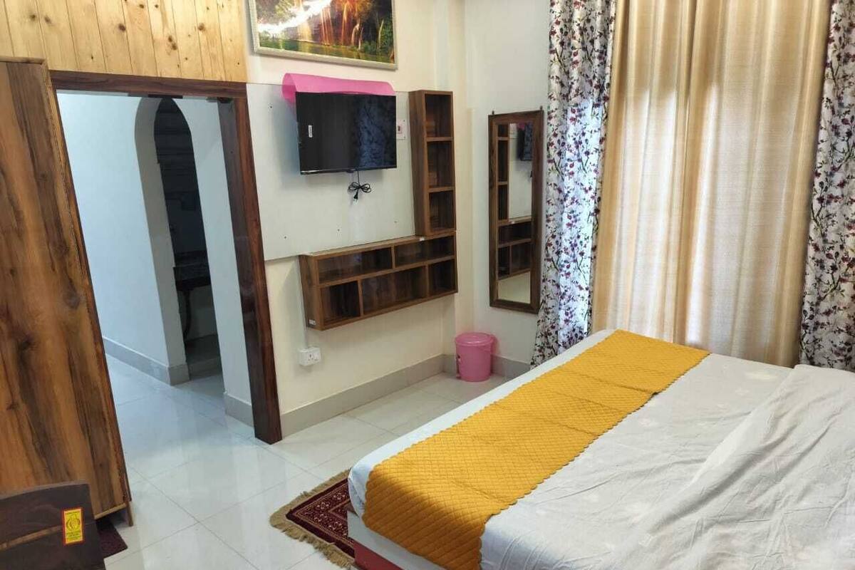Un dormitorio con una cama, un televisor montado en la pared, estantes de madera, un cubo de basura rosa y una cortina estampada con flores.