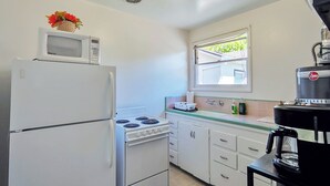Une cuisine compacte avec un réfrigérateur blanc, une cuisinière et un four à micro-ondes. Il y a une fenêtre donnant sur un espace verdoyant, un évier avec un plan de travail et une machine à café posée sur une petite table.