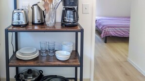 Un coin cuisine avec une étagère en bois sur laquelle sont posés une machine à café, un grille-pain, une bouilloire et une boîte de café. Sous l’étagère, on aperçoit des assiettes, des verres et une poêle antiadhésive. On distingue également un lit recouvert d’une couverture à rayures à l’arrière-plan.
