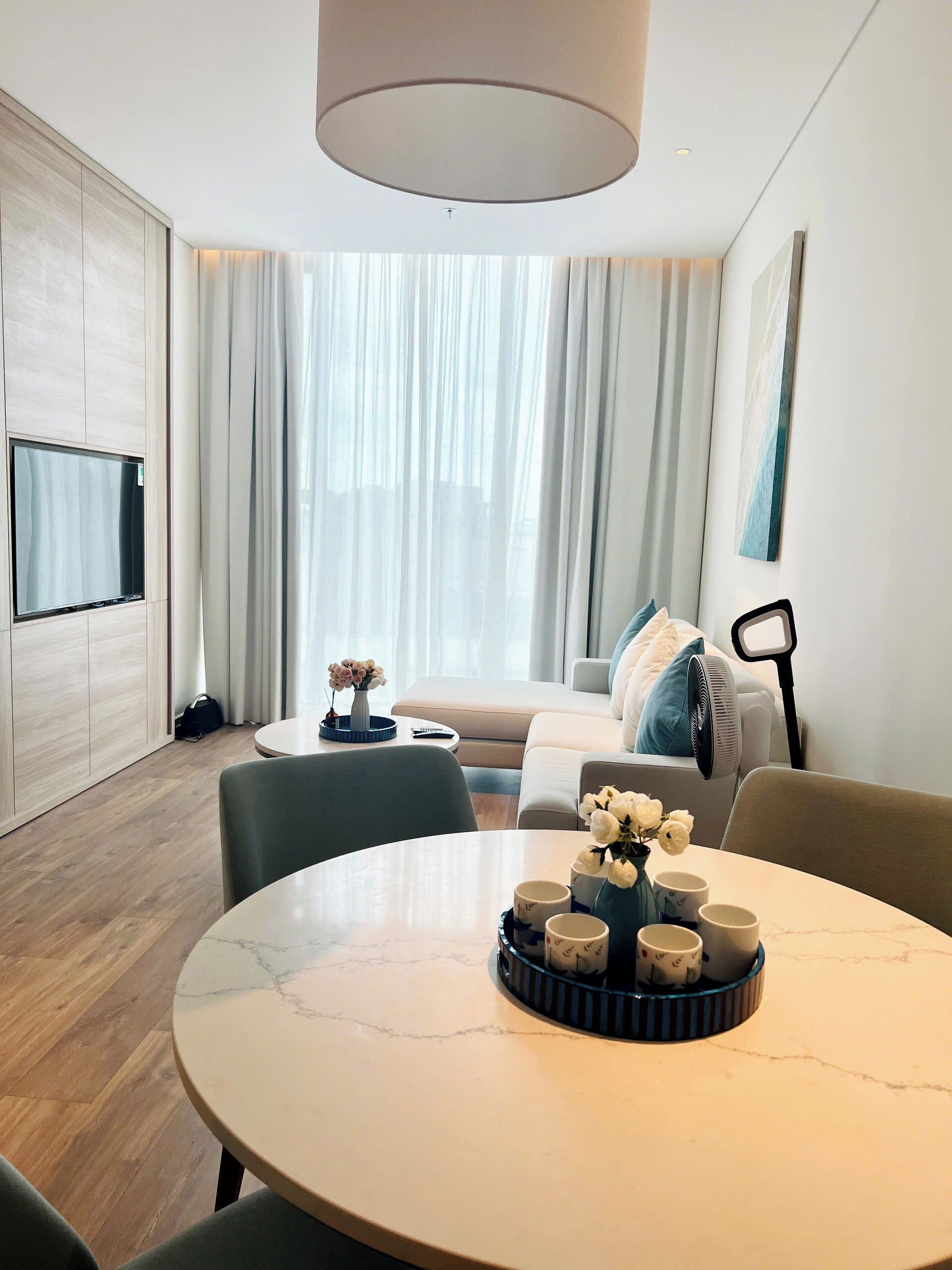 A modern hotel room with a round marble table, a sofa, a TV mounted on the wall, and a large window with curtains.