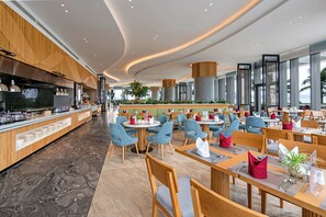 A modern restaurant with a long counter, round tables, and blue chairs. - Alacarte Ha Long Bay Managed by Navis Global (Bai Chay)