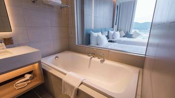 A modern bathroom with a large bathtub, a vanity with a mirror, and a view of a bedroom with a bed and pillows.
