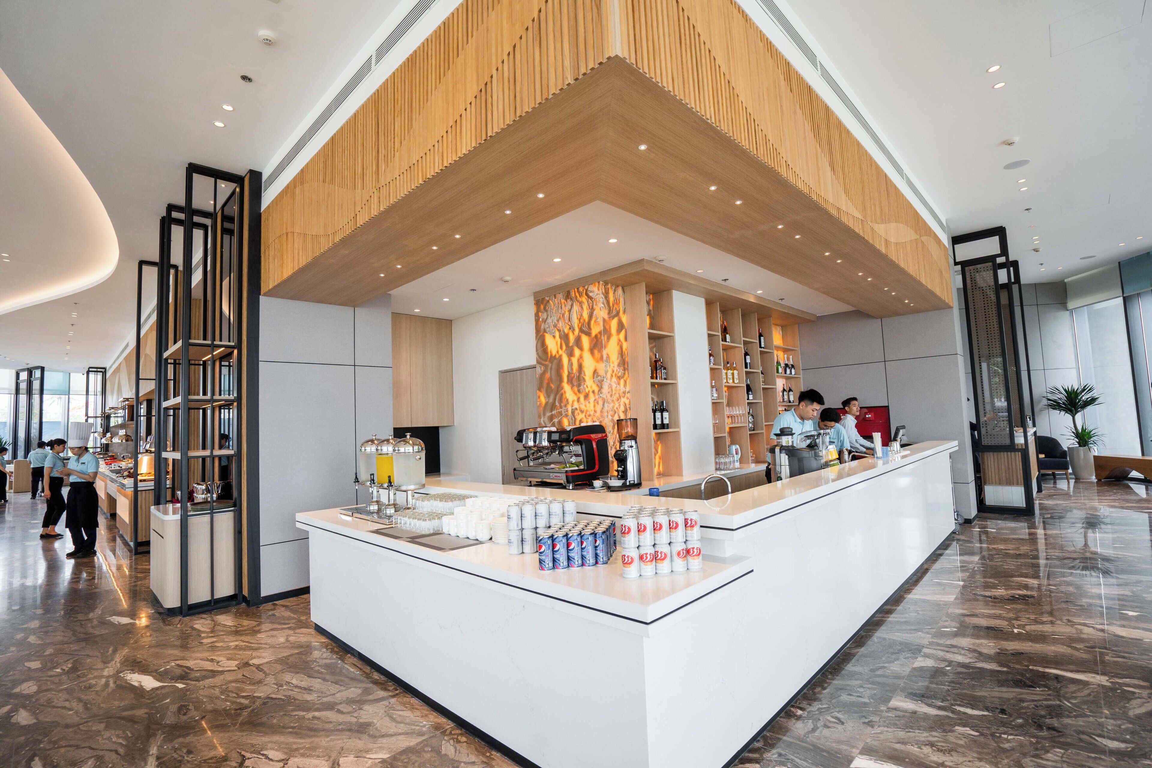 A modern hotel lobby with a central reception desk, a coffee bar, and a seating area with a large plant.