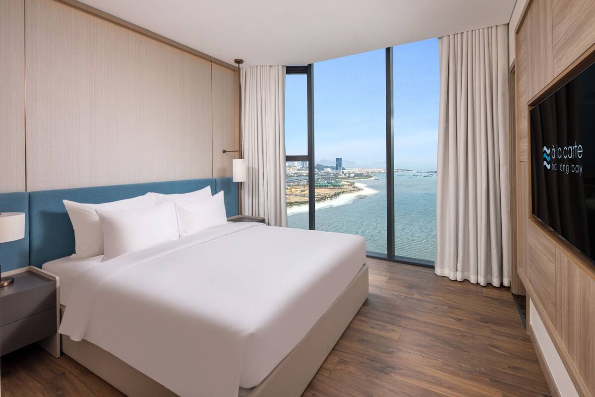 A hotel room with a large bed, a nightstand, a TV, and a view of a coastal city.