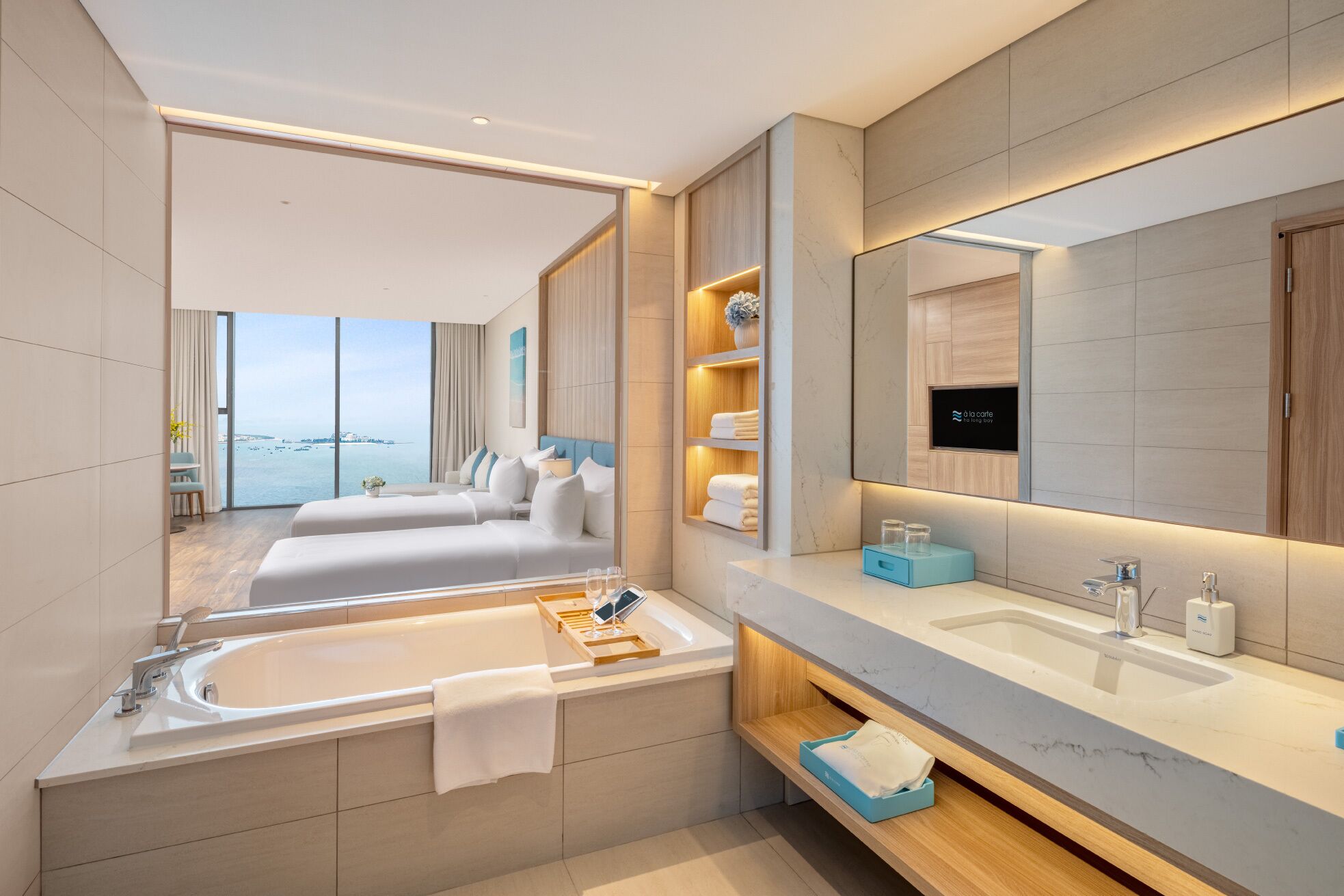 A modern bathroom with a large bathtub, two sinks, and a vanity. There is a large mirror above the vanity and a view of the sea through the window.