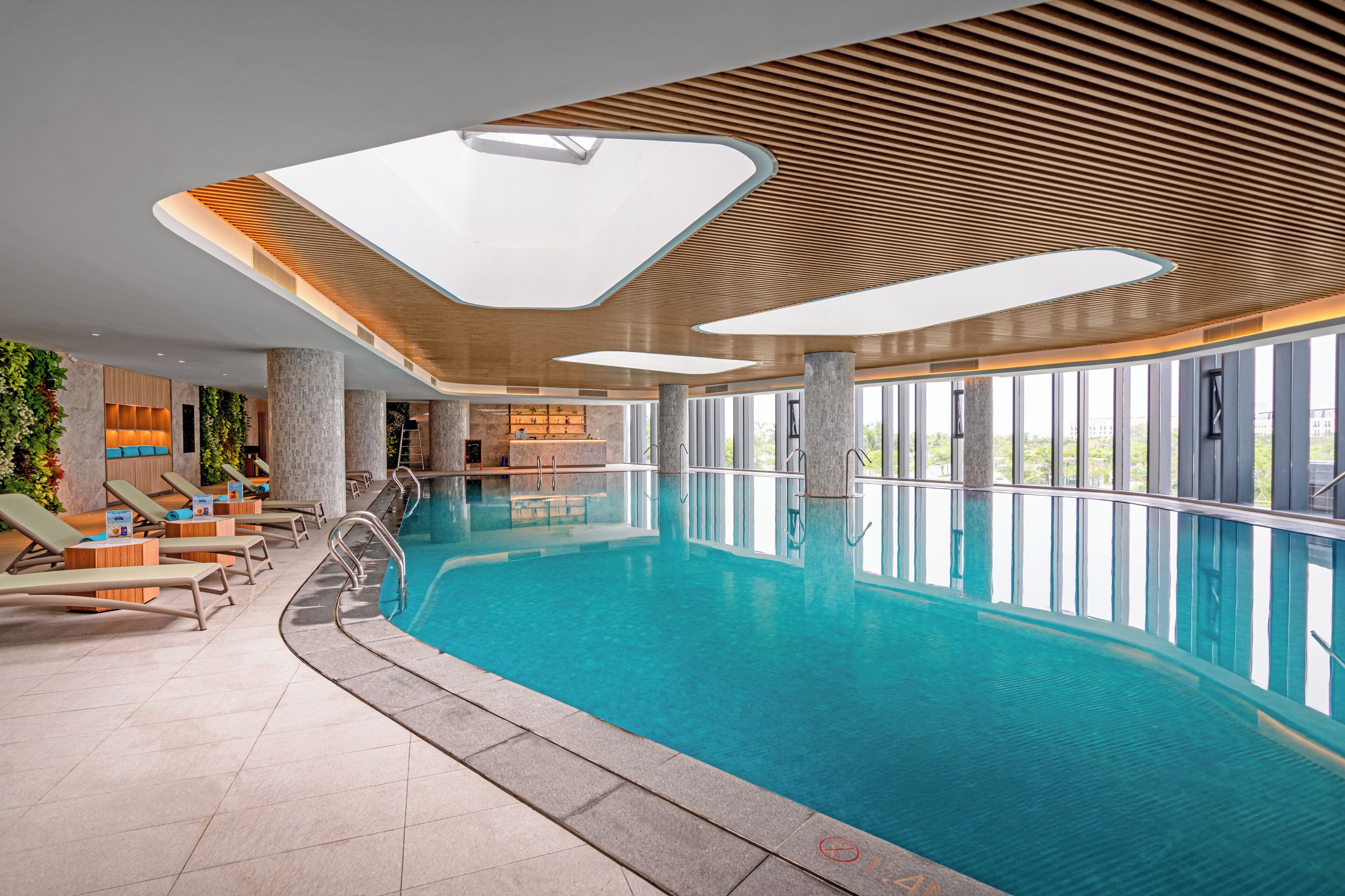An indoor swimming pool with lounge chairs, large windows, and a modern ceiling design.