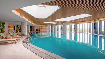 An indoor swimming pool with lounge chairs, large windows, and a modern ceiling design.