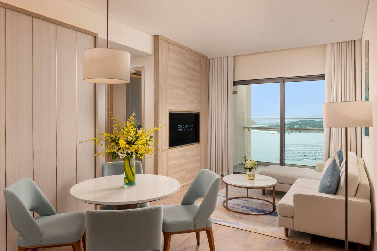 A modern hotel room with a dining area, a sofa, and a large window offering a view of the sea.